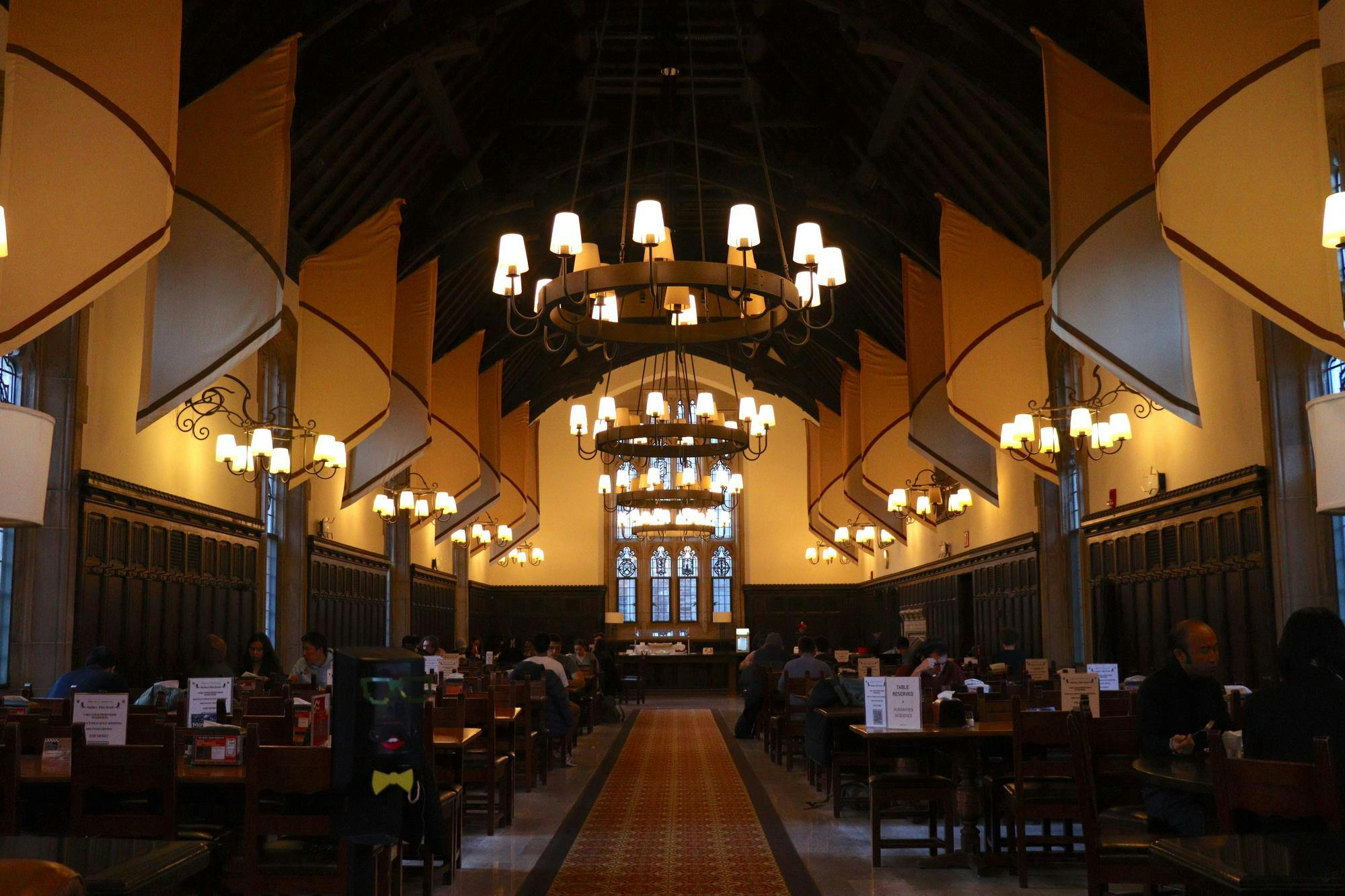 A chandelier hangs from the center of the room, which other small chandeliers on the side. A dark red rug is in the middle of the room with multiple long wooden tables with wooden chairs on each side.