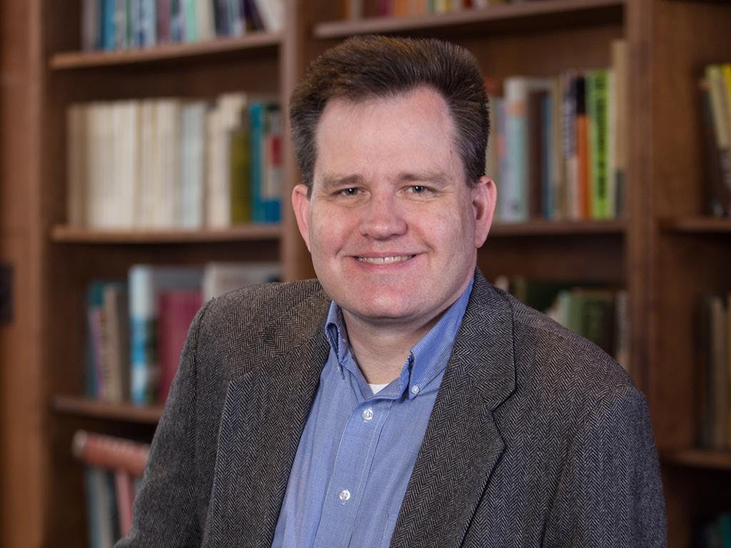 A photo of Keith Whittington wearing a grey suit and blue dress shirt. He is smiling. In the background, there are full bookshelves. 