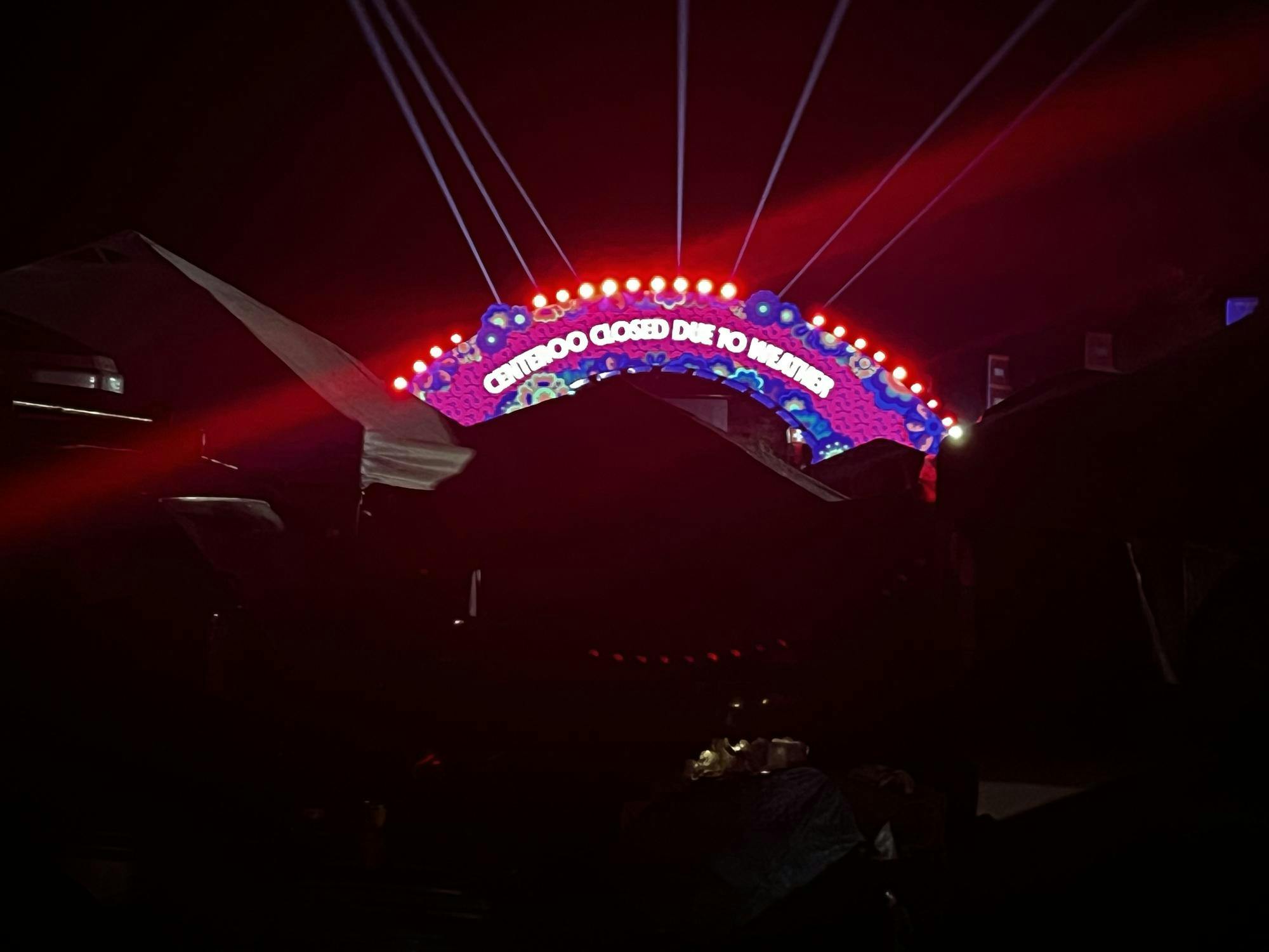 A large pink and purple arch with electronic signage displays the message “Centeroo closed due to weather.” The arch is captured in surrounding darkness with silhouettes of white tents and temporary structures surrounding it.