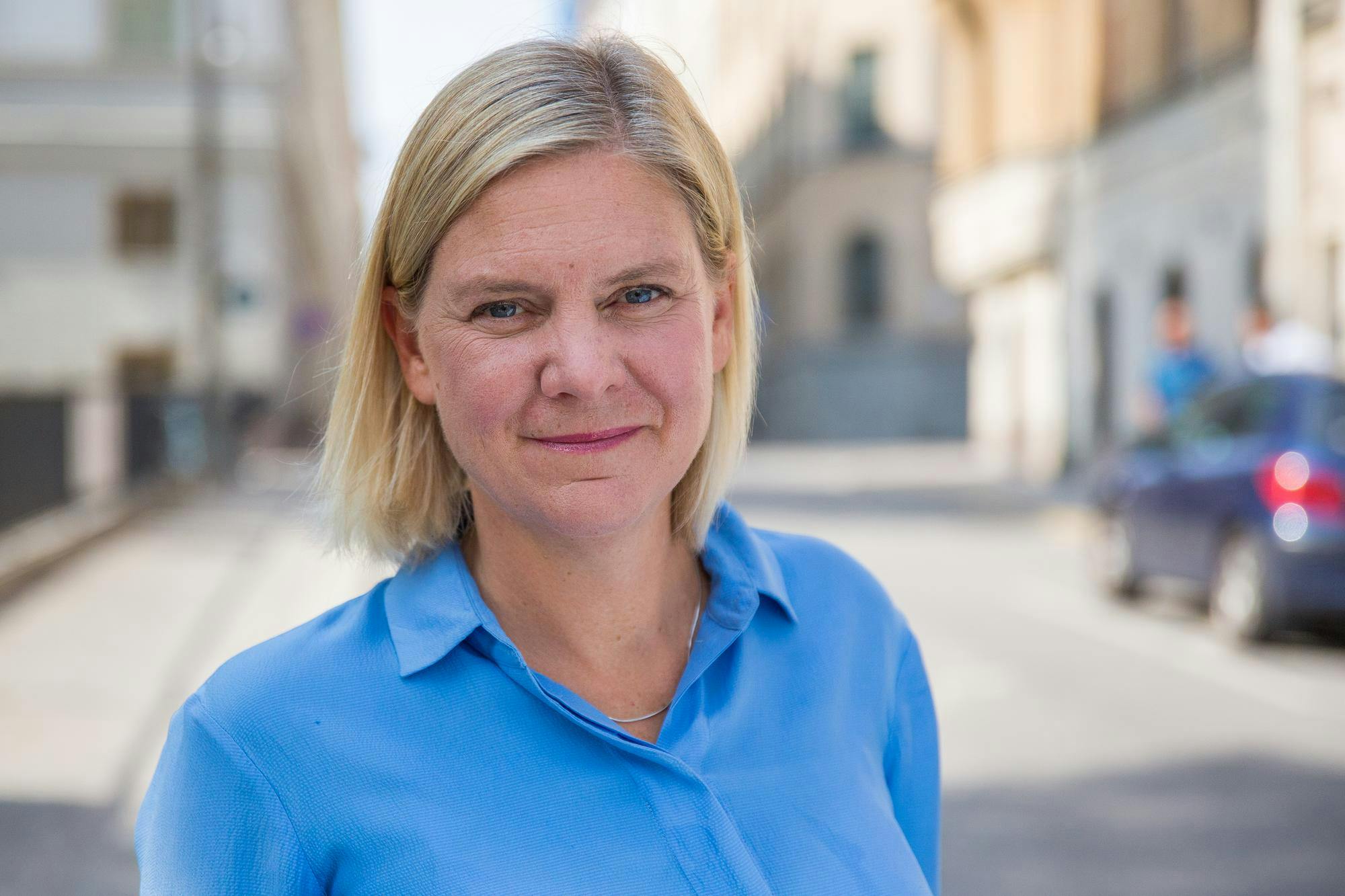 middle-aged woman with blond hair and blue eyes with a terse smile wearing a blue shirt in front of a blurred urban background with a car in it.