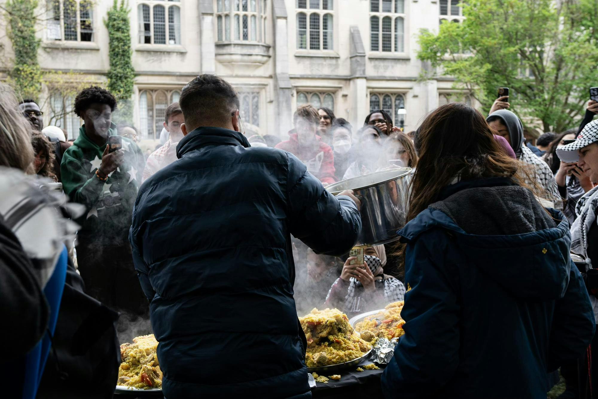 A crowd of protestors is gathered around a table as several organizers serve maqluba for a meal. Many people also have their phones out to take pictures and videos of the food.