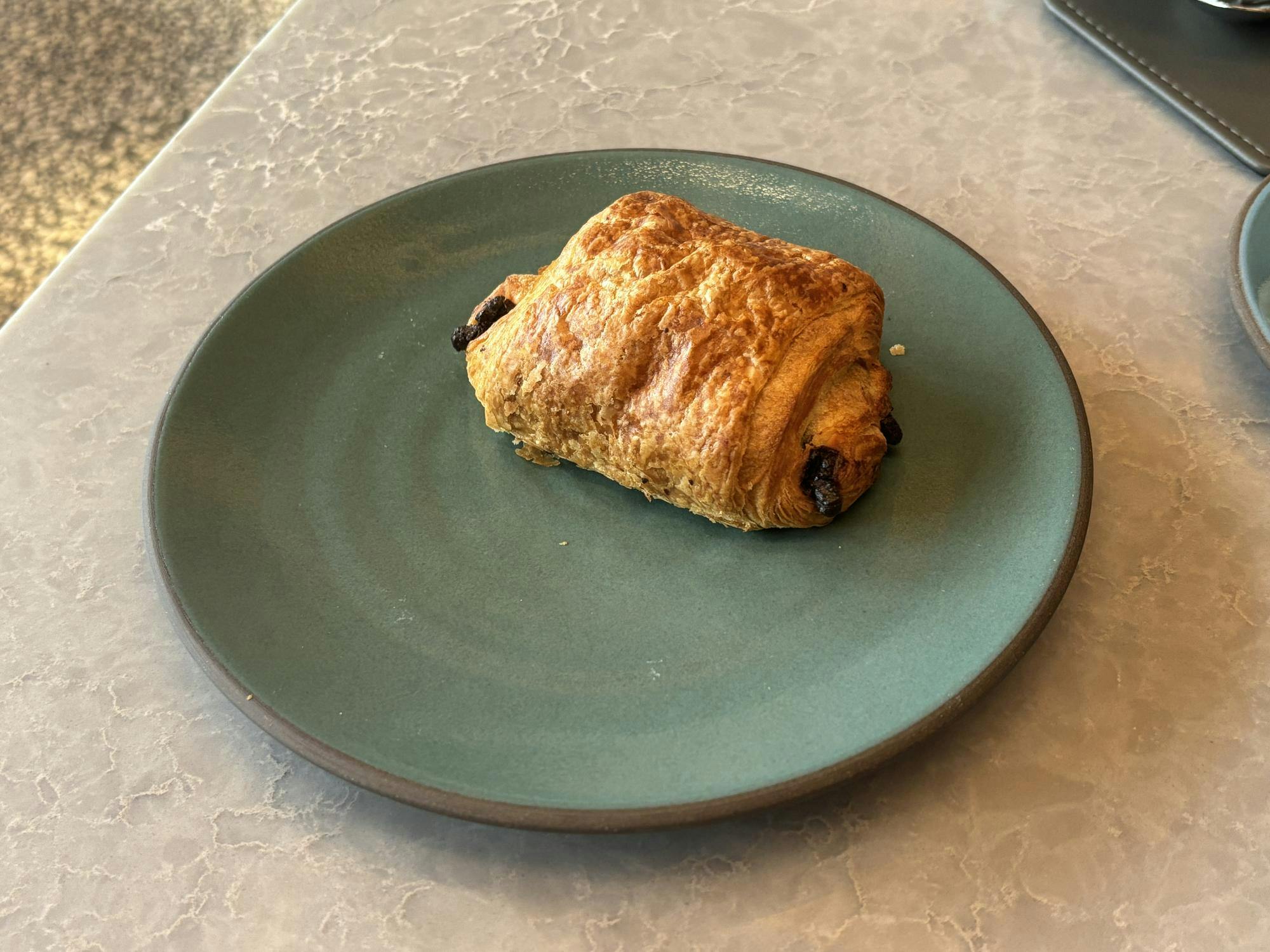 A light brown roll of croissant has brown chocolate sticking out the ends. It sits on a teal plate.