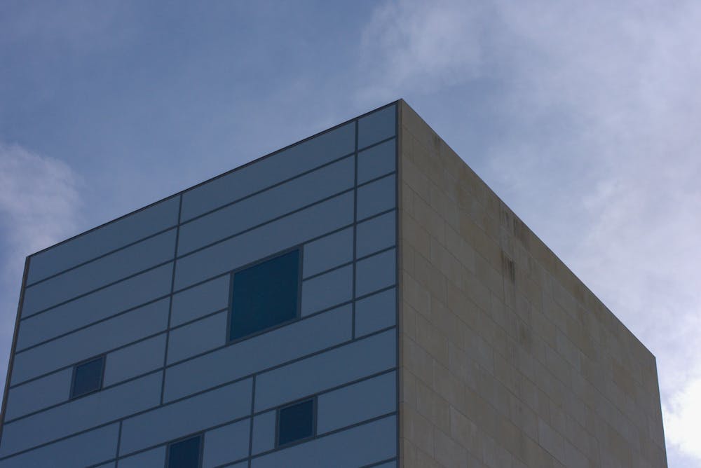 The gray Lewis Center for the Arts building sits below a blue sky checkered with white clouds. Half of the building is solid concrete with no windows and the other half is glass that reflects the blue sky.