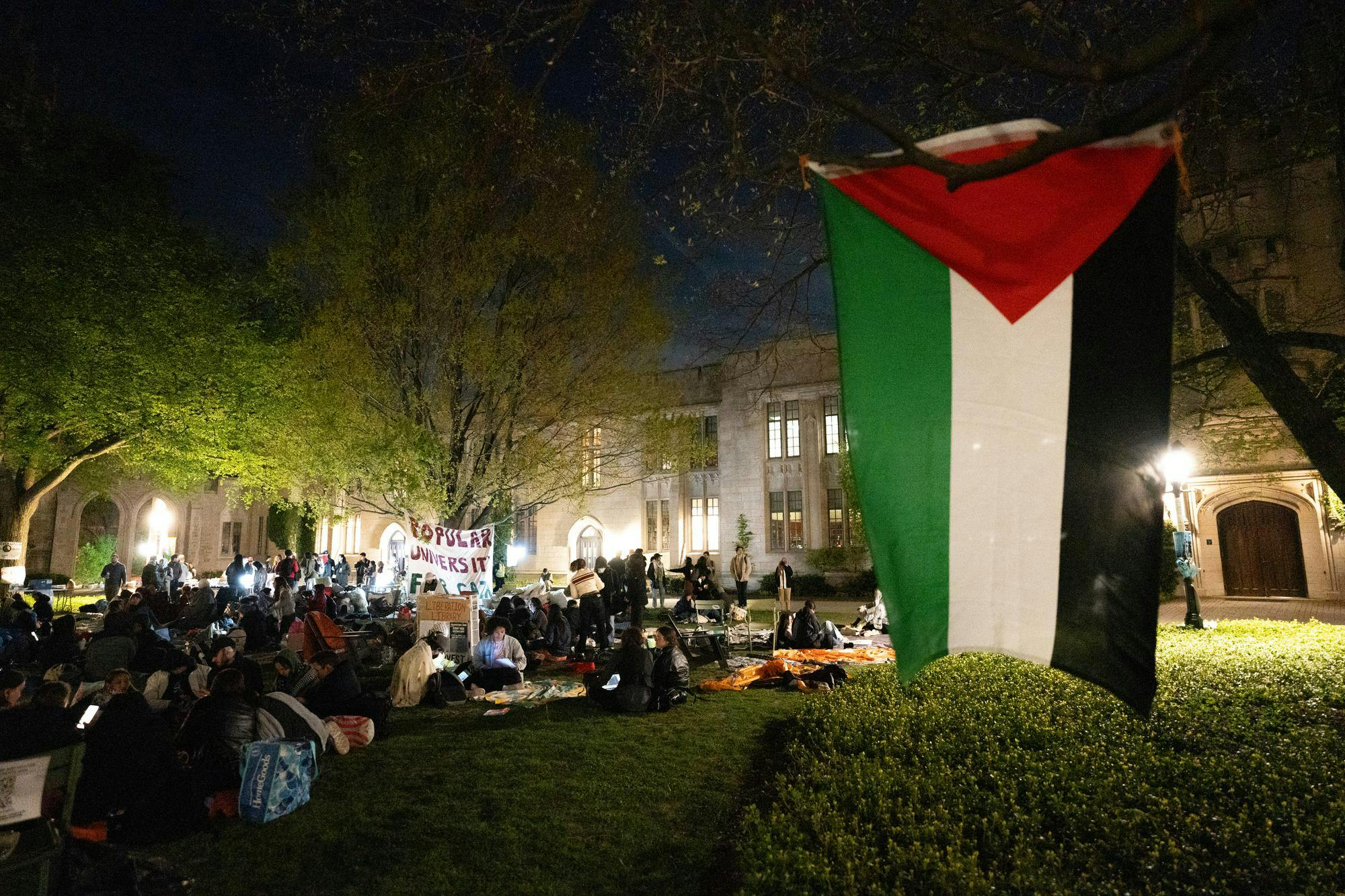 Students sit on a lawn in the dark. In the foreground, a flag with green, white, black, and red hangs from a tree. 