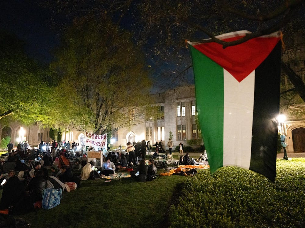 Students sit on a lawn in the dark. In the foreground, a flag with green, white, black, and red hangs from a tree. 