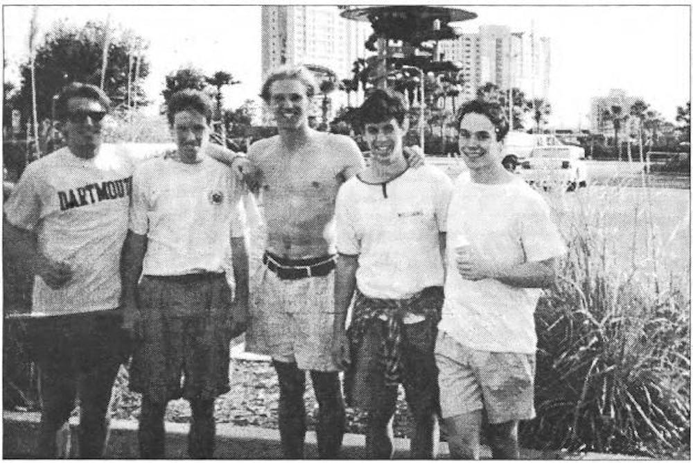 A black and white photograph of a line of 5 men smiling at the camera, wearing summer attire.