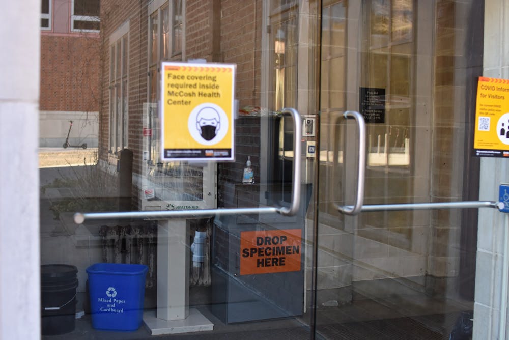 Glasss door with sign "Face covering required inside McCosh health center" with metal box with words "Drop Specimen Here" inside.