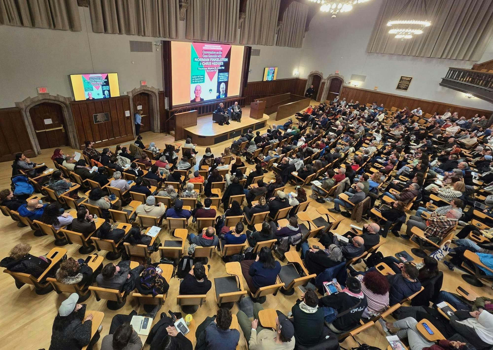 An overhead photograph of a filled auditorium, with two speakers sitting on stage.