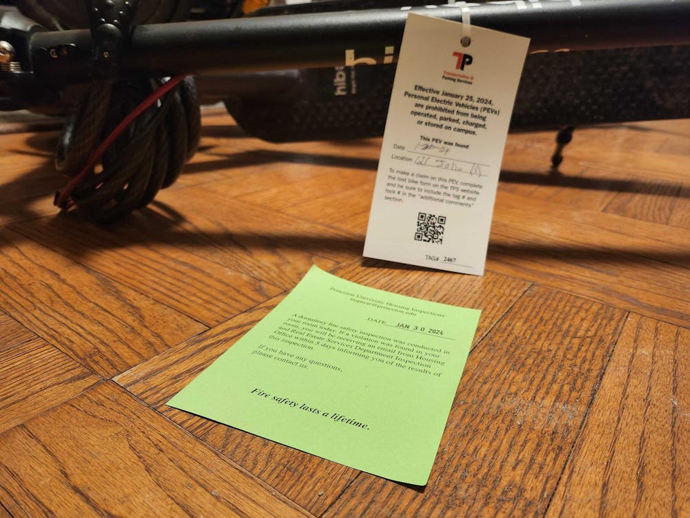 An electric scooter on wooden flooring, tagged with a reminder of the personal electric vehicle (PEV) policy. A green slip of paper in front of the scooter, confirming the dormitory received a fire inspection on Jan. 30 2024.
