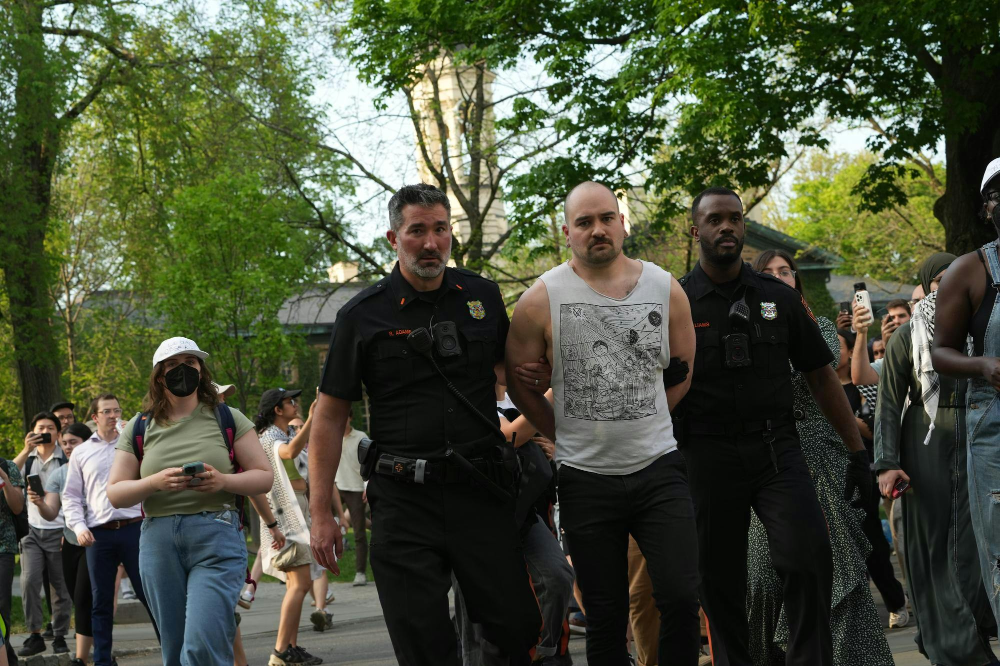 A man in a white graphic muscle tee walks with his hands behind his back while two police officers grab each of his arms in black and orange uniforms. Many people stand around them.
