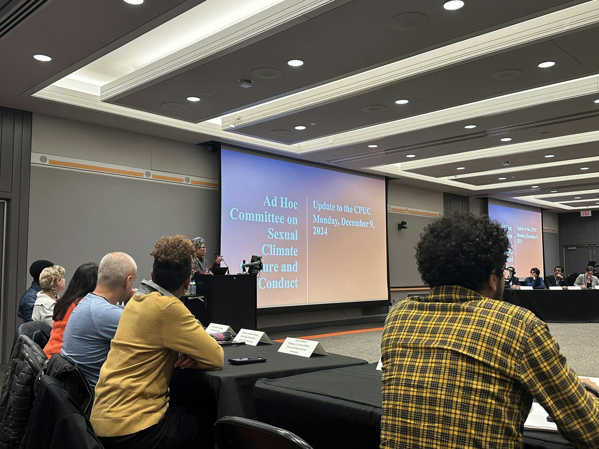 A conference room with attendees seated around a table and facing a large screen at the front. The screen displays a presentation titled "Ad Hoc Committee on Sexual Climate, Culture, and Conduct: Update to the CPUC, Monday, December 9, 2024." A speaker stands at a podium to the left of the screen, addressing the group. 