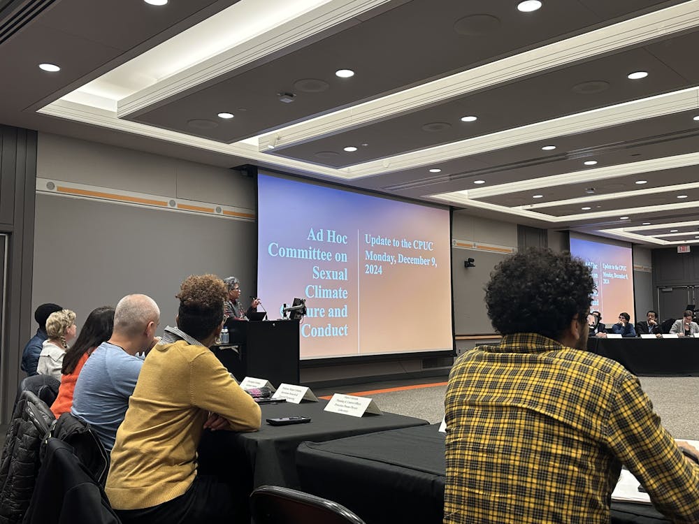A conference room with attendees seated around a table and facing a large screen at the front. The screen displays a presentation titled "Ad Hoc Committee on Sexual Climate, Culture, and Conduct: Update to the CPUC, Monday, December 9, 2024." A speaker stands at a podium to the left of the screen, addressing the group.