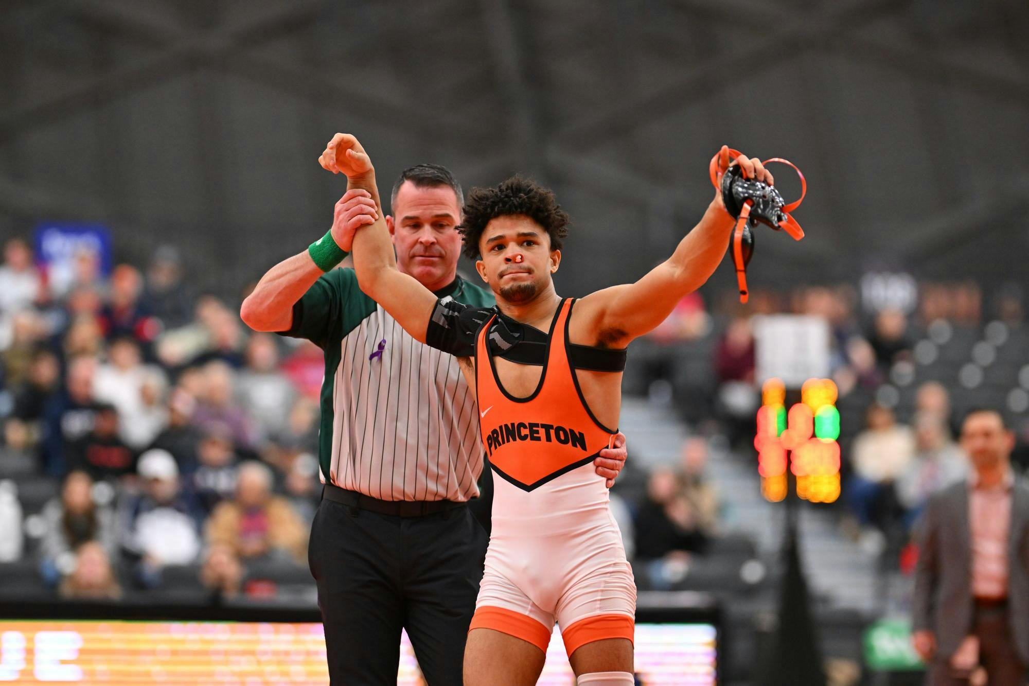 Man in orange and black wrestling leotard with tissue stuffed in a bloody nose has his hand raised victoriously by an official in grey and black pinstripes.