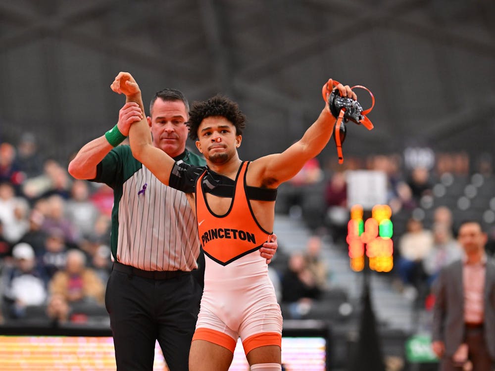 Man in orange and black wrestling leotard with tissue stuffed in a bloody nose has his hand raised victoriously by an official in grey and black pinstripes.