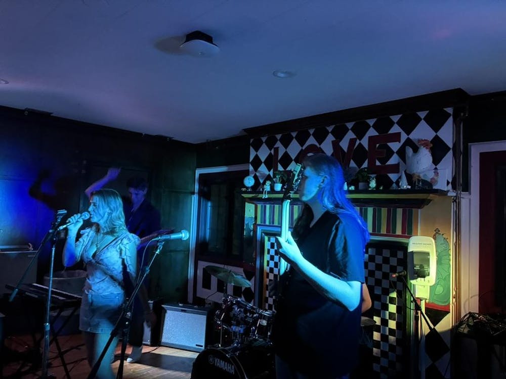 Three individuals perform and play music in front of a checkerboard fireplace in a dimly-lit room. 