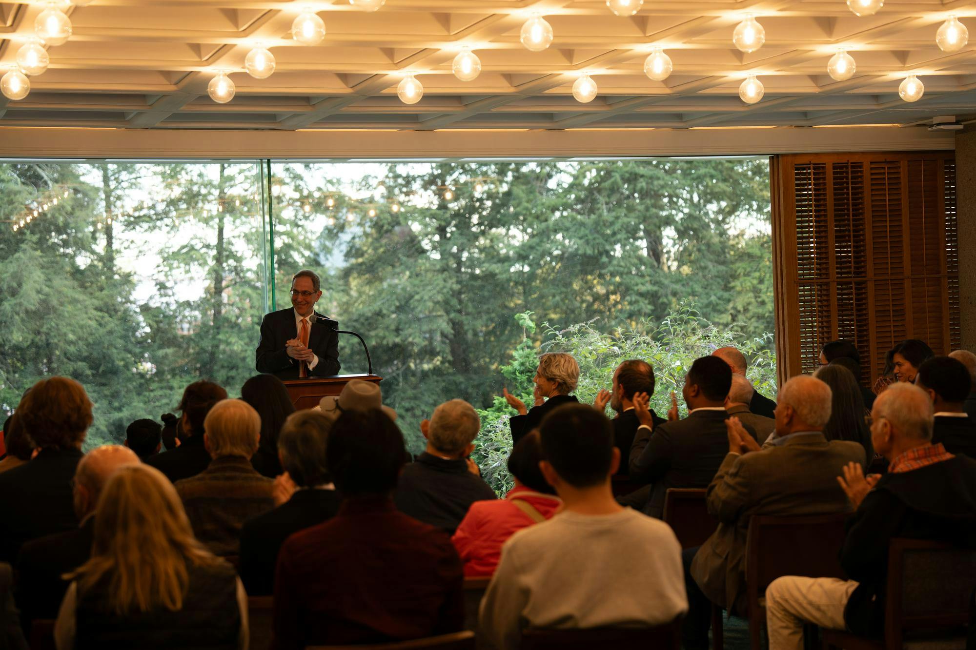 A man on a podium clapping while the audience in chairs also claps. 