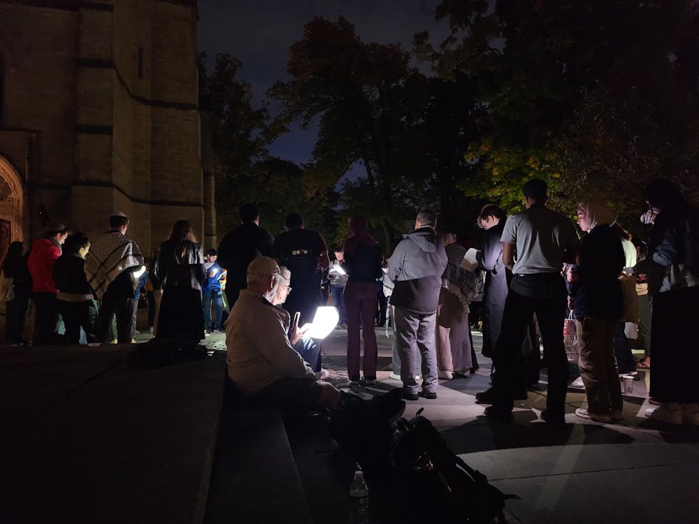 A group of people gather in a circle in the background. The lighting is dim. In the foreground an elderly couple is sitting and looking down at a light.