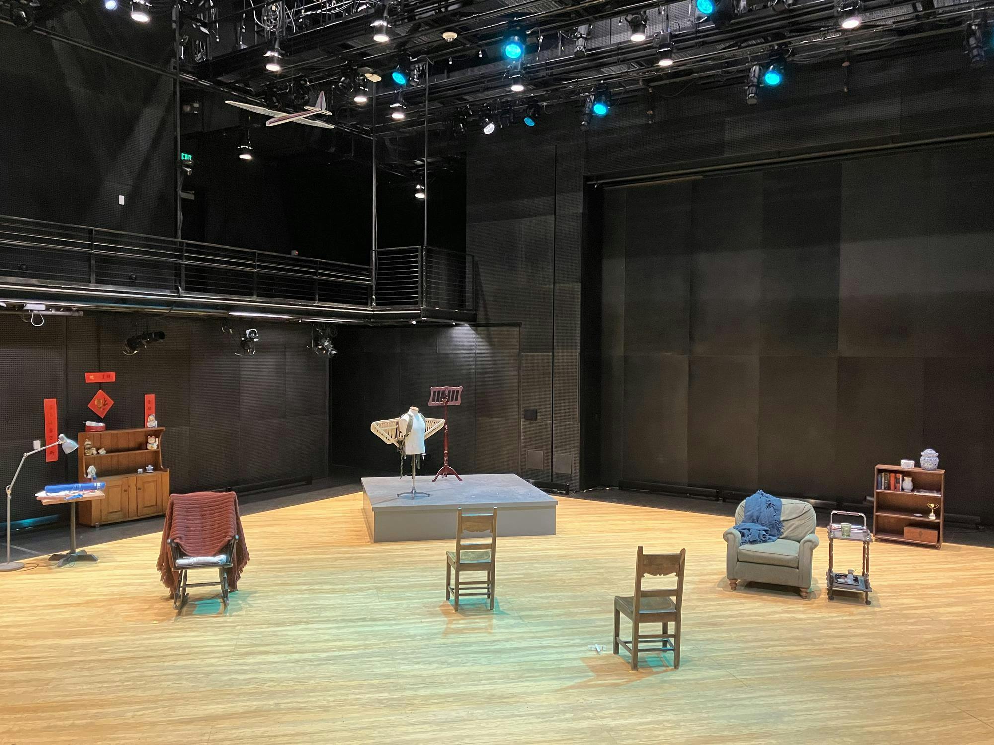 A wooden stage is set with wooden chairs, a gray sofa chair, and an elevated platform that has a mannequin wearing wings and a music stand on it.