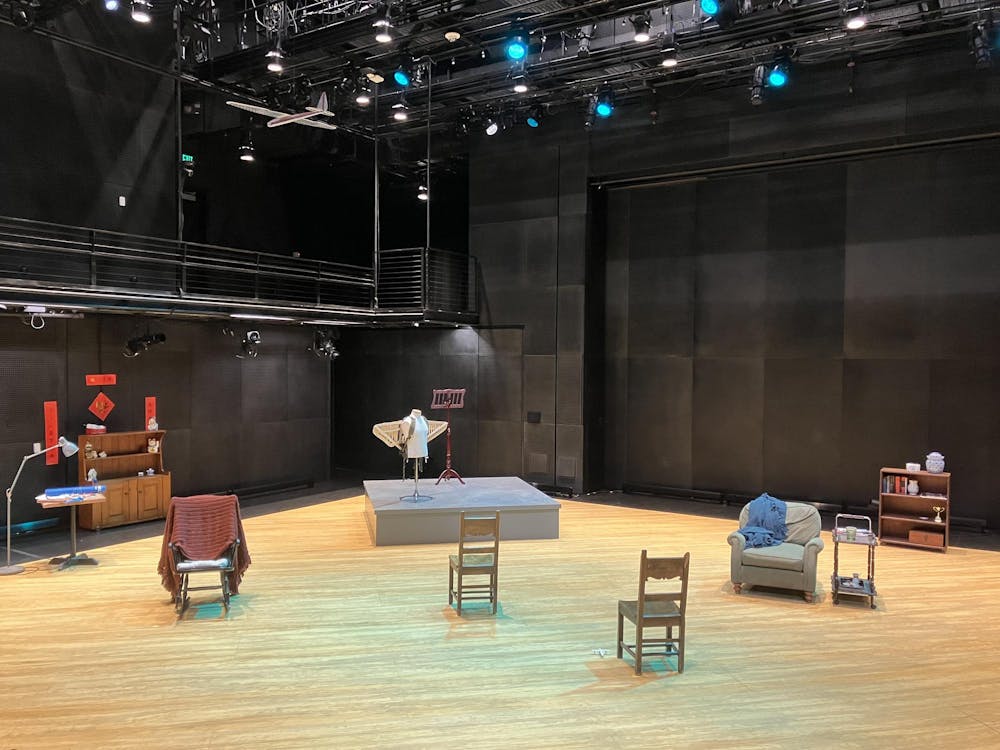 A wooden stage is set with wooden chairs, a gray sofa chair, and an elevated platform that has a mannequin wearing wings and a music stand on it.