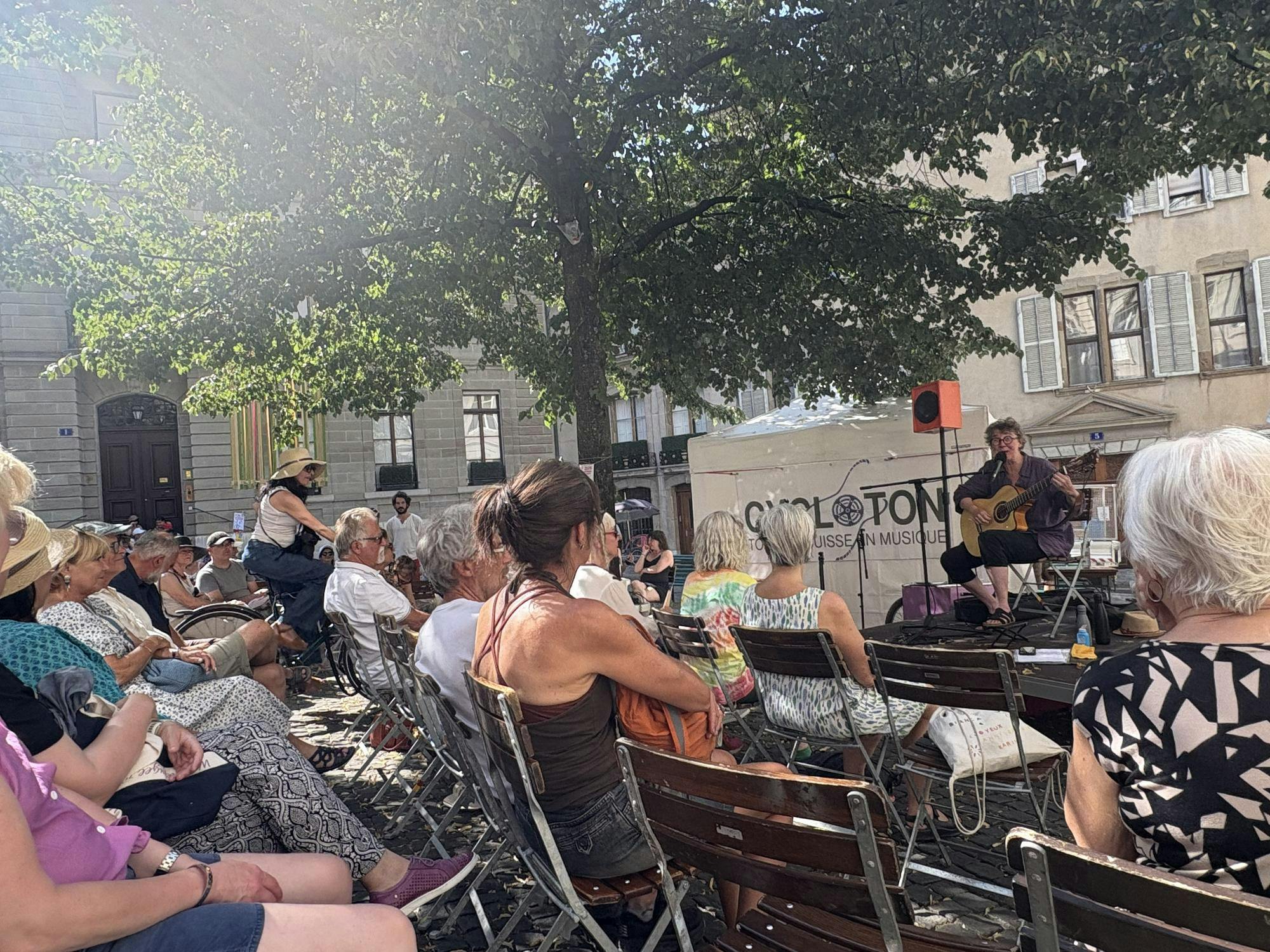 A group of people are sitting in brown wooden chairs. The people are wearing clothing of various colors and styles. You can see the backs of the people as they are all facing a stage on the right side of the image. On the stage, an adult person is wearing a purple shirt, black capri style pants, black glasses, and black sandals. The person is seated; they appear to be singing into a microphone and playing a tan-colored acoustic guitar.