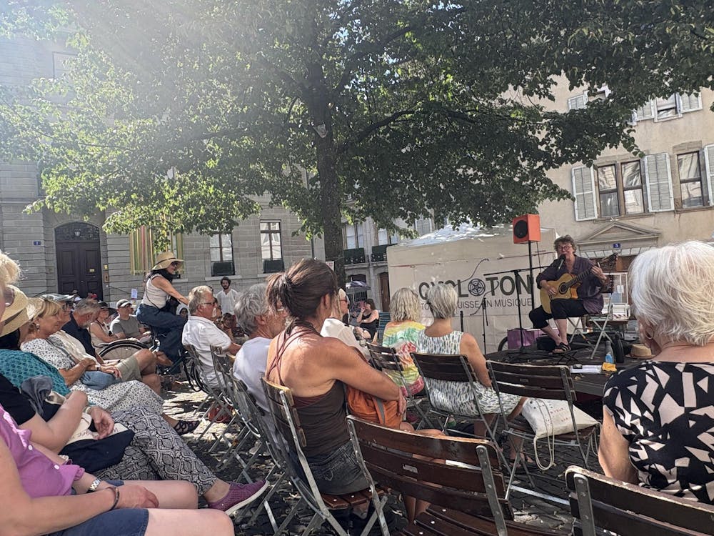 A group of people are sitting in brown wooden chairs. The people are wearing clothing of various colors and styles. You can see the backs of the people as they are all facing a stage on the right side of the image. On the stage, an adult person is wearing a purple shirt, black capri style pants, black glasses, and black sandals. The person is seated; they appear to be singing into a microphone and playing a tan-colored acoustic guitar.