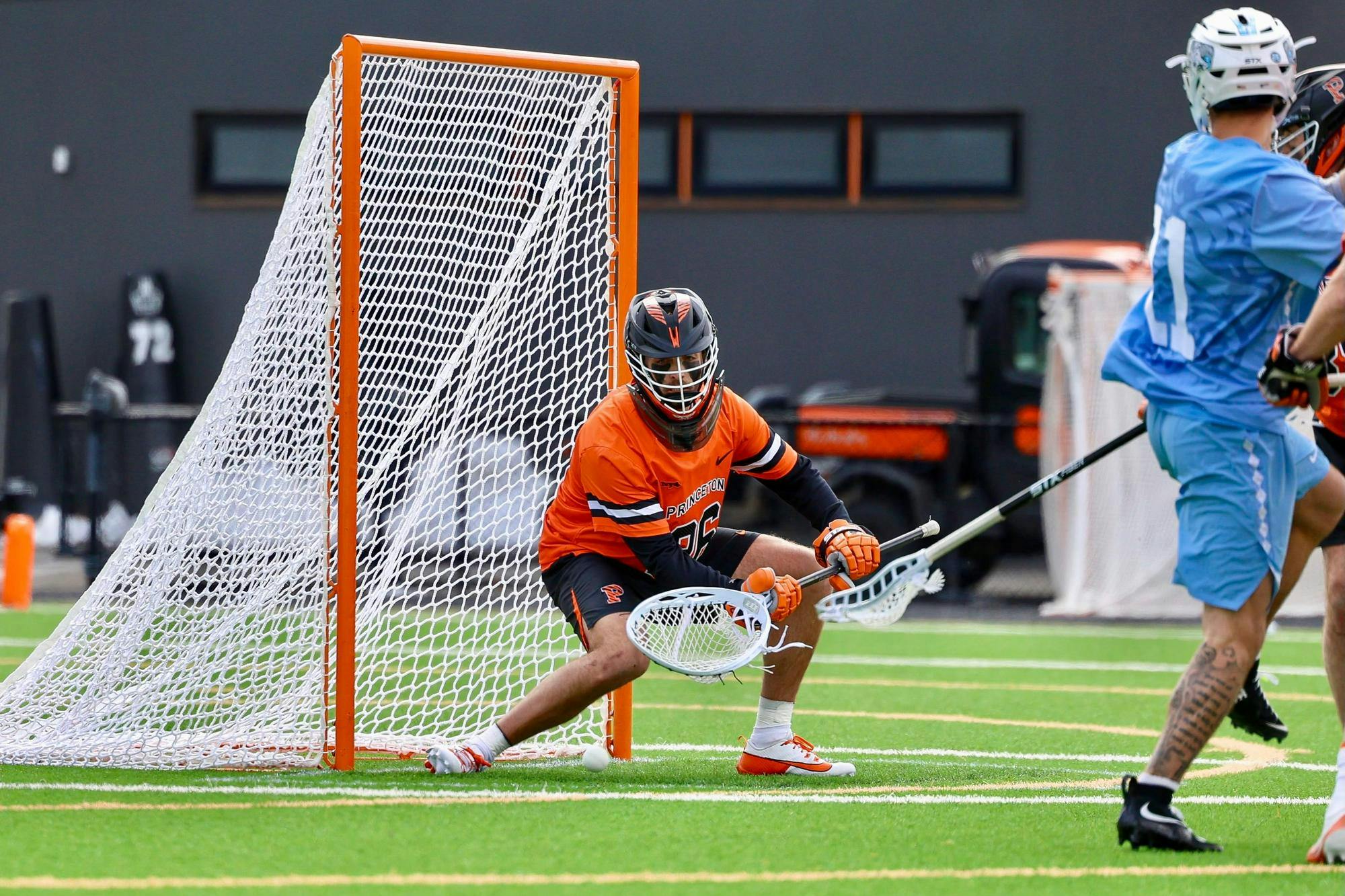 A Princeton lacrosse goalie in orange and black makes a low save in front of the net as a North Carolina player in light blue attacks.