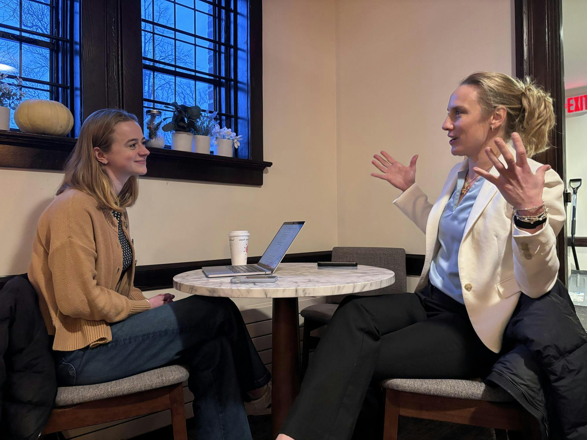 A woman in a brown sweater talks to a woman in a white blazer and blue top, who is talking with her hands. 