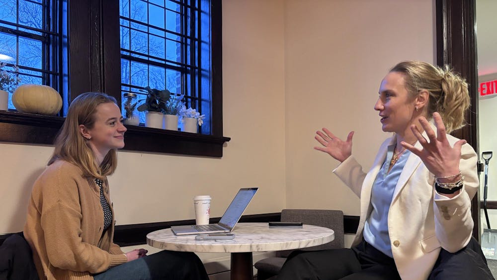A woman in a brown sweater talks to a woman in a white blazer and blue top, who is talking with her hands.
