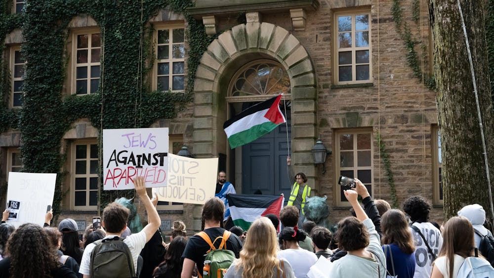 A crowd of students gathers in front of a large brick building covered with ivy. The students hold signs and raise their fists up in the air. One student in the front of the crowd holds a Palestinian flag up in the air.