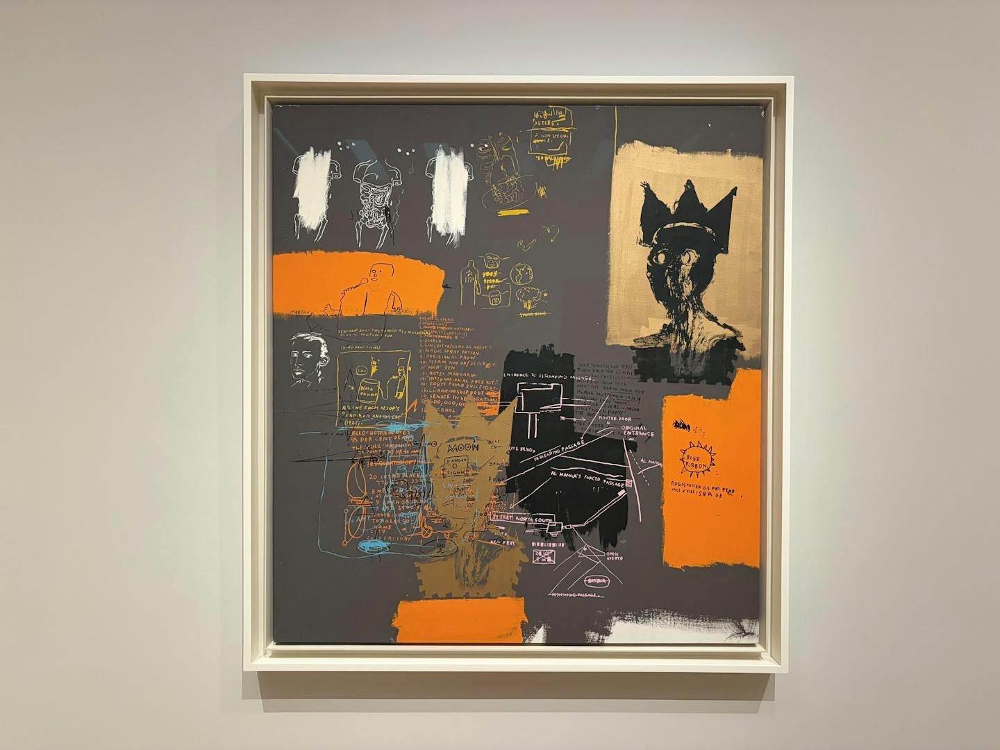 A painting with a dark grey background, sketches of anatomical models, random phrases, and a black silhouette with a crown hangs against a white wall.
