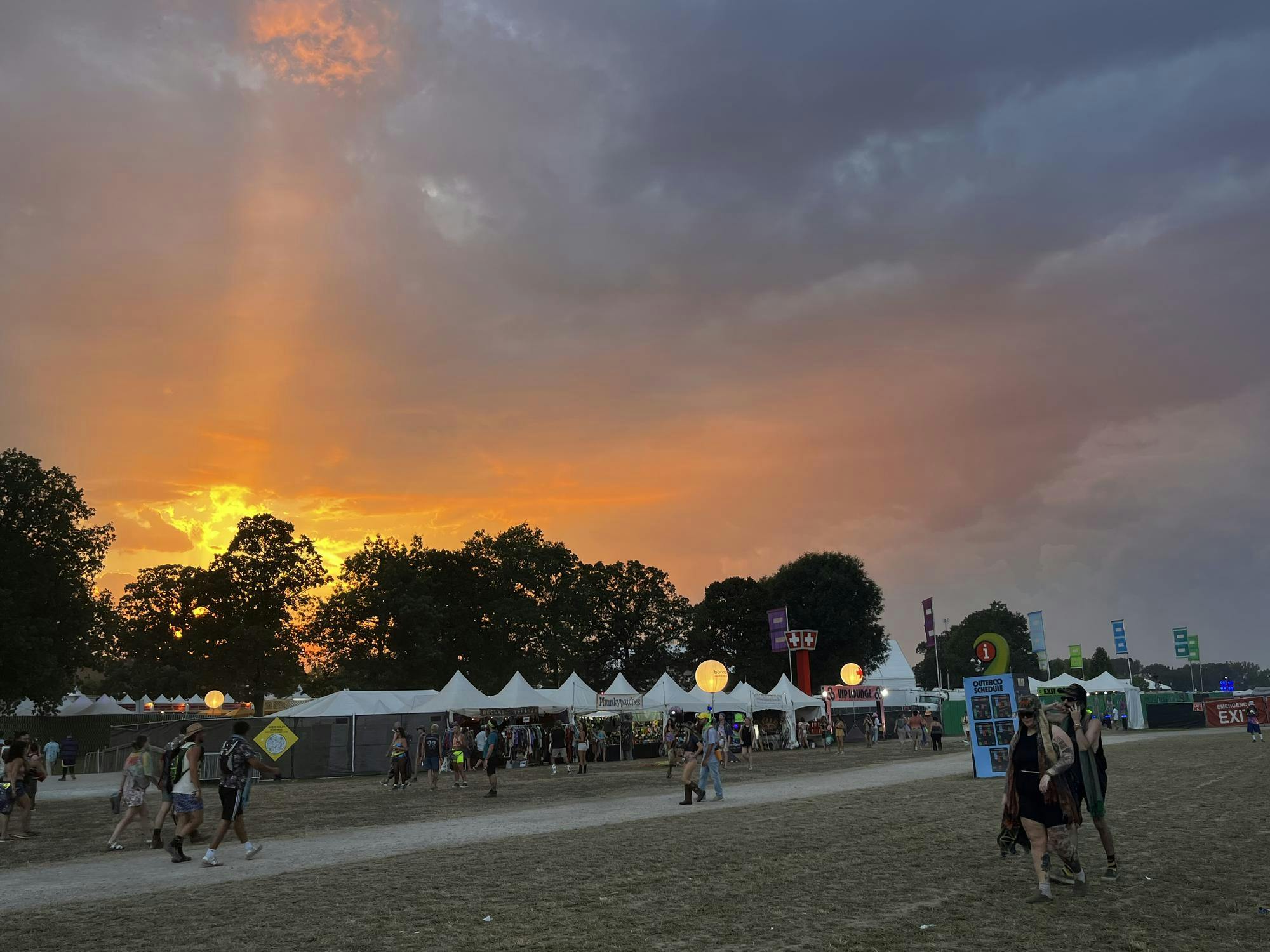 Attendees of the Bonnaroo music festival stroll before a series of white tents as the sun sets over a the silhouettes of trees.