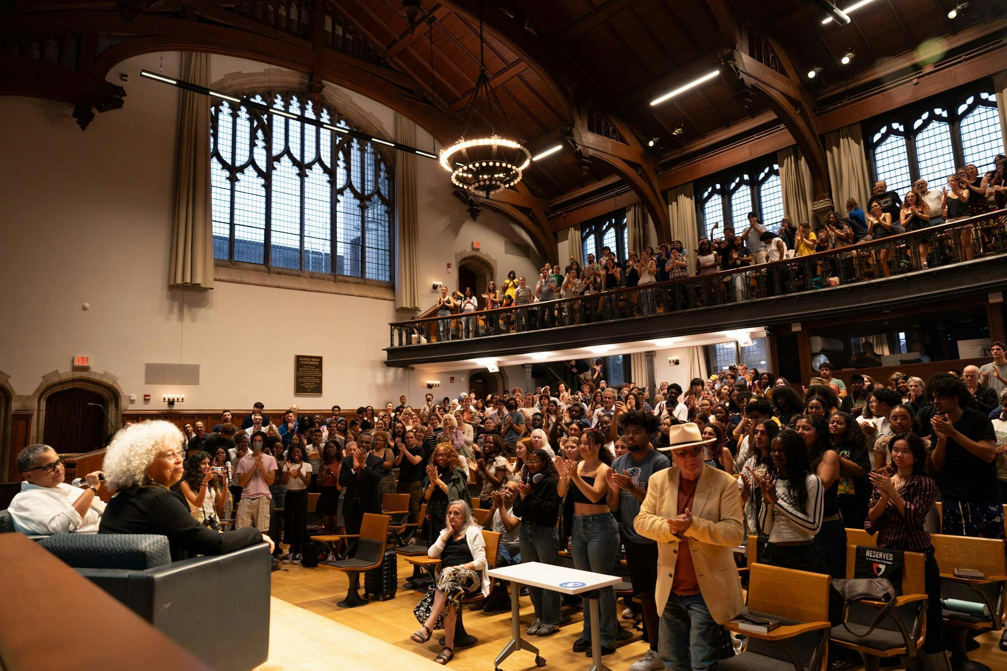 A large audience of about 400 people stands and applauds in a cavernous room with a ground floor and upper observation deck. In the lower left corner, a speaker and a moderator, both women, are seated on a stage. 