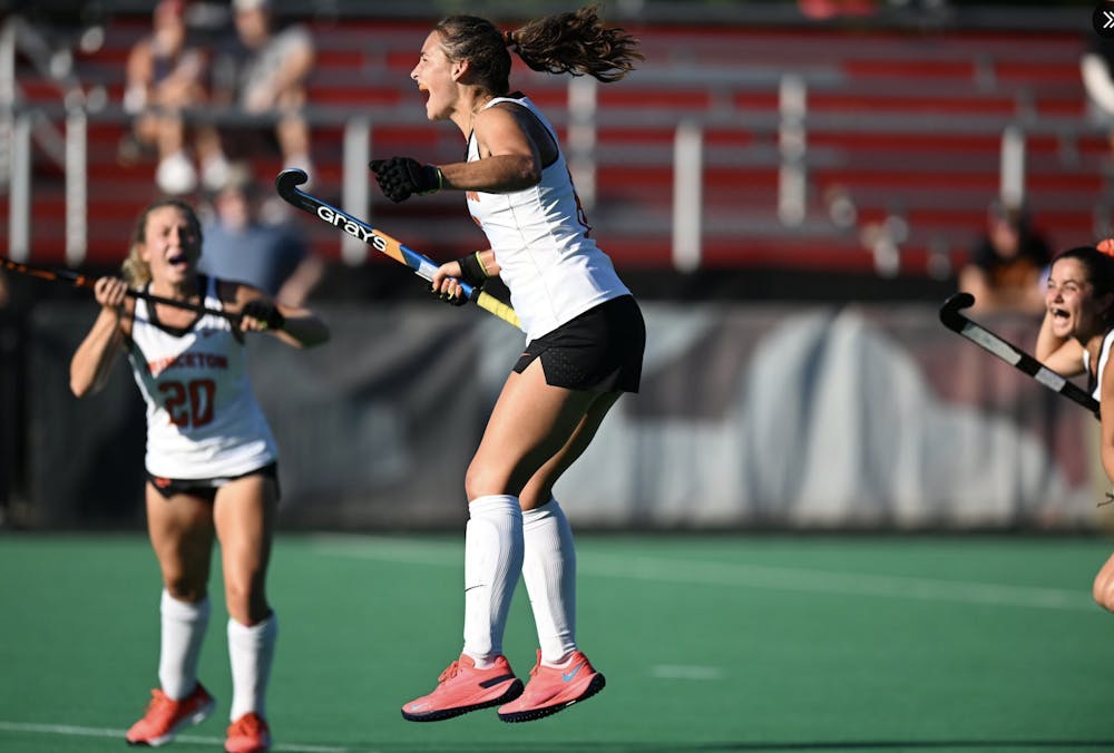 A field hockey player wearing a white jersey jumps celebrating with a stick. Players also wearing white jerseys celebrate in the background.  