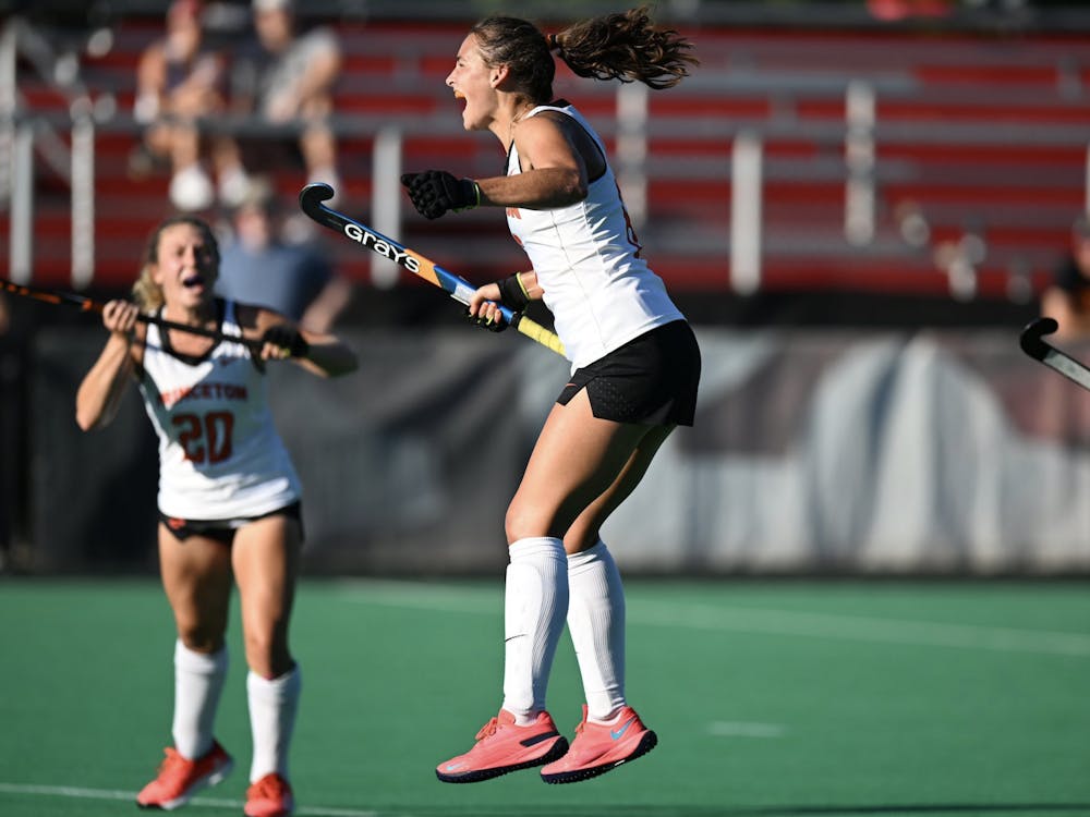 A field hockey player wearing a white jersey jumps celebrating with a stick. Players also wearing white jerseys celebrate in the background.  