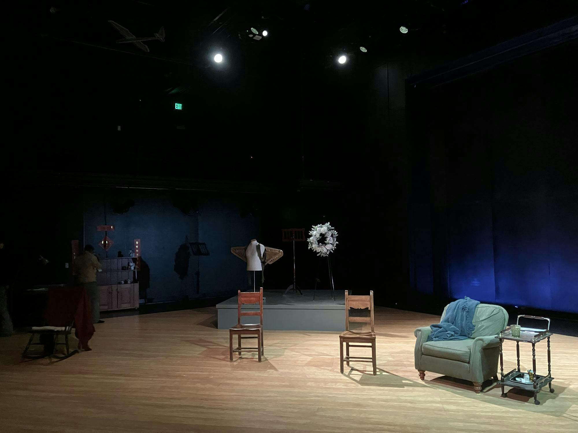 
A wooden stage with dimmed lights is set with wooden chairs, a gray sofa chair, and an elevated platform that has a mannequin wearing wings and a music stand on it.