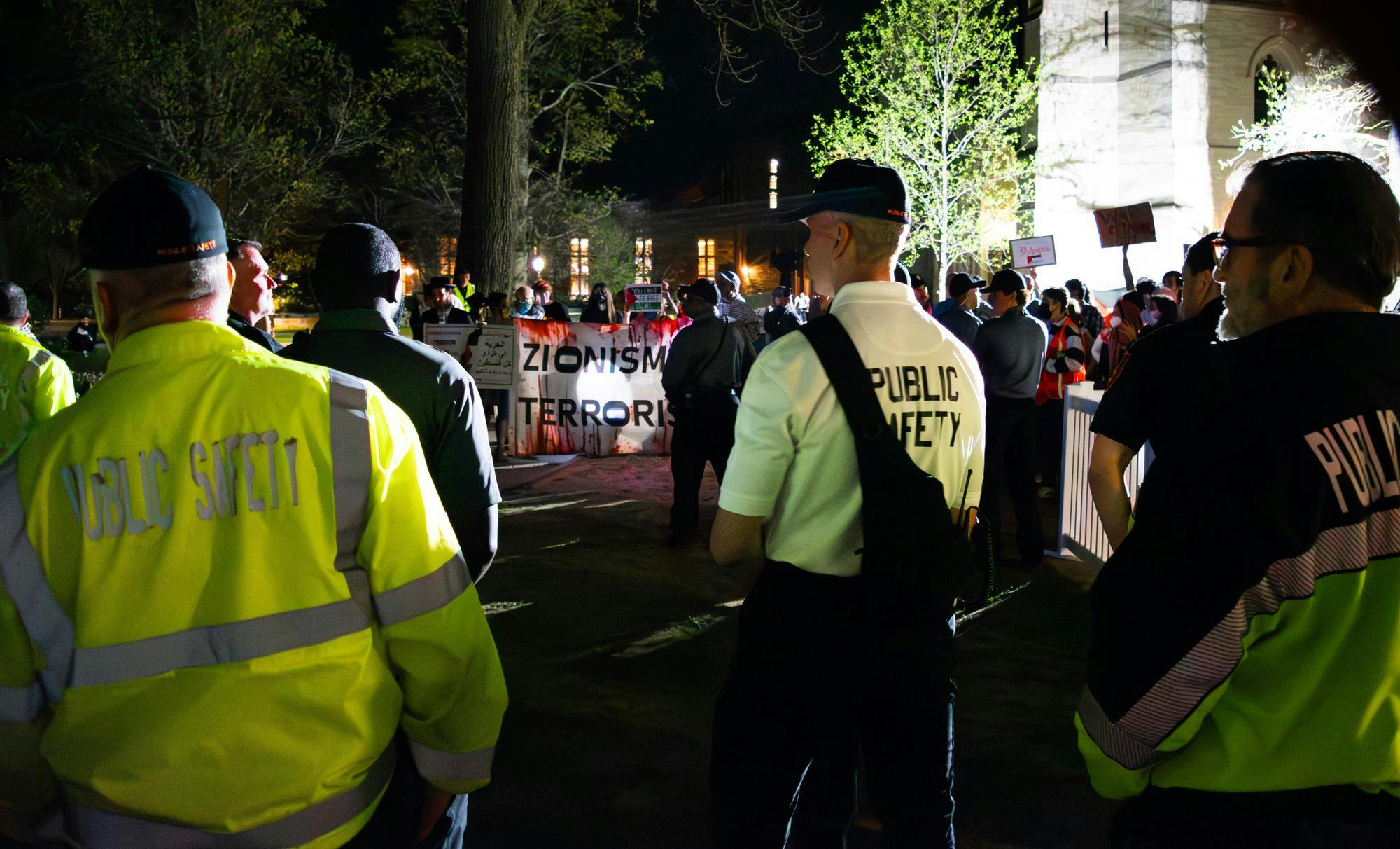 Three people with vests and shirts reading "public safety" stand across from a sign reading "Zionism Terrorism"