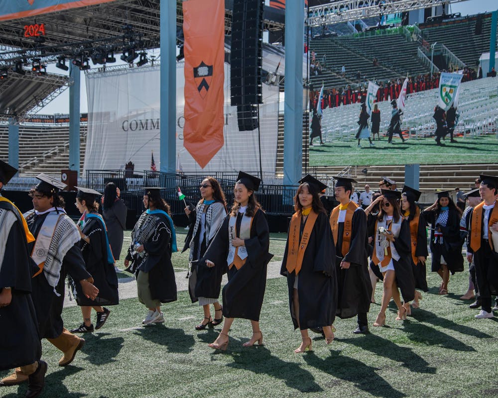 Graduates and black robes walk across a green field with a stage in the background.