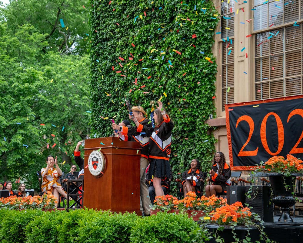 Three students wearing class day jackets pop confetti behind a Princeton podium.
