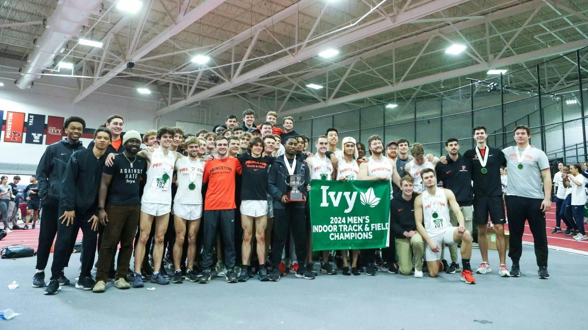 Group of men stand huddled together holding banner reading "Ivy 2024 Men's Indoor Track and Field Championships" by an indoor track.