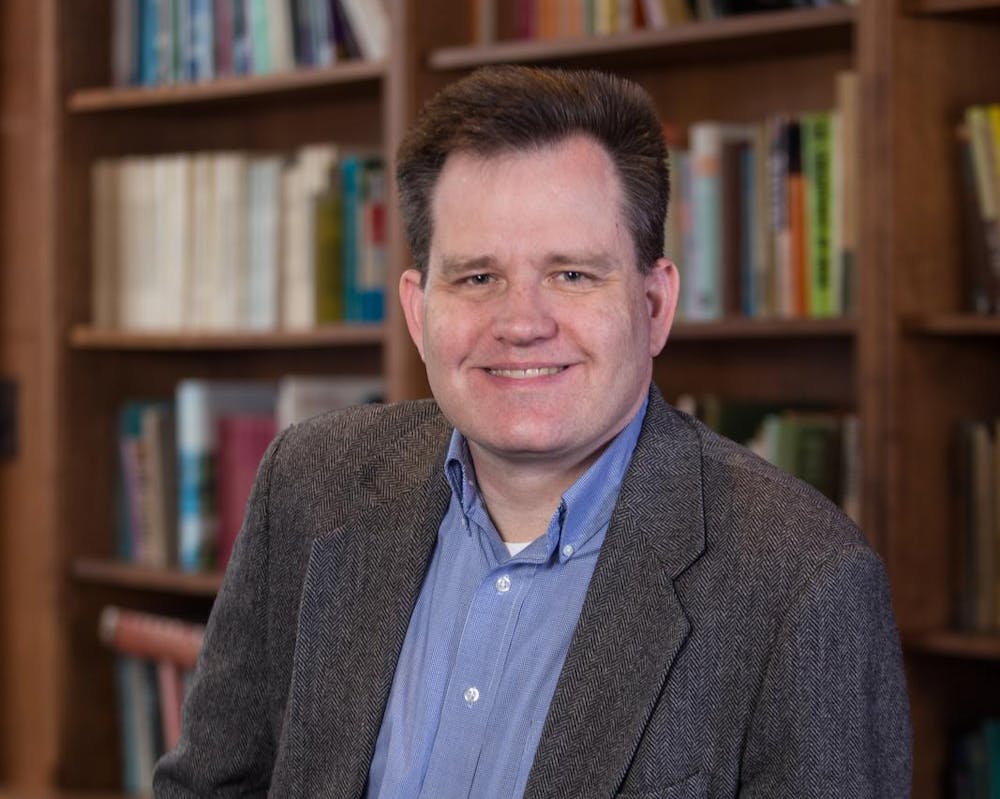 A photo of Keith Whittington wearing a grey suit and blue dress shirt. He is smiling. In the background, there are full bookshelves. 