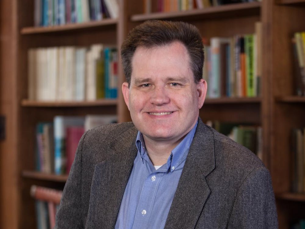 A photo of Keith Whittington wearing a grey suit and blue dress shirt. He is smiling. In the background, there are full bookshelves. 