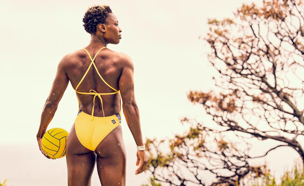 Woman in water polo bathing suit stands with ball at her side, facing away from camera 