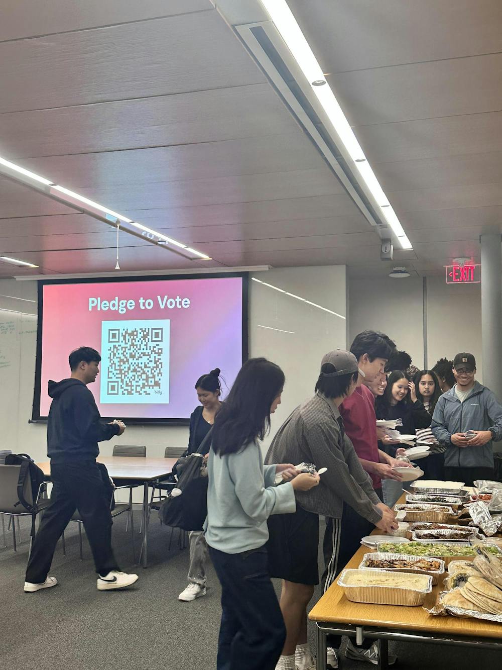 A group of young adults get food out of tin containers on a table. A screen reading “Pledge to Vote” with a QR code is in the center of the image.