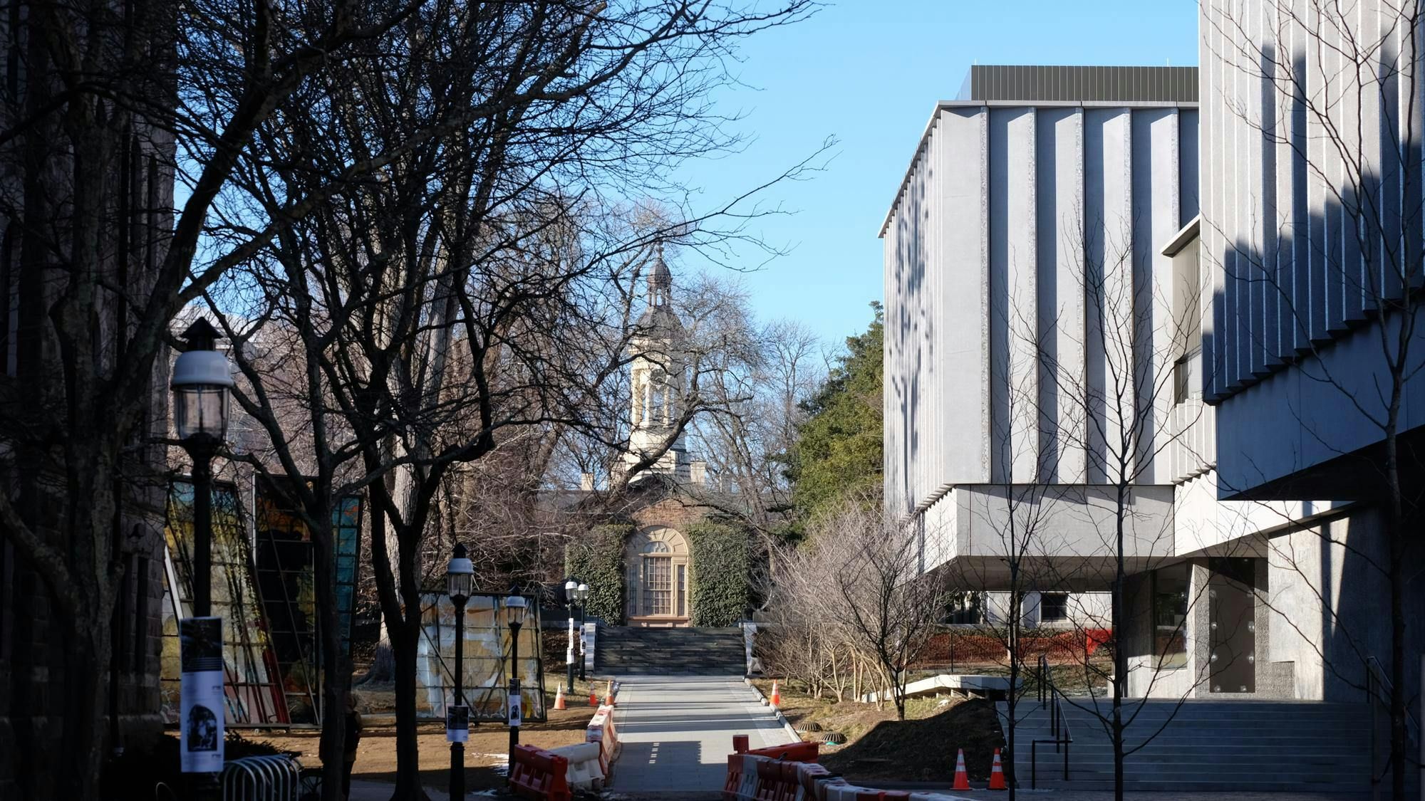 A path leading up to Nassau Hall lined with construction cones and barriers with the Art Museum's concrete exterior on the right and a few bare trees and lamp posts on the left.