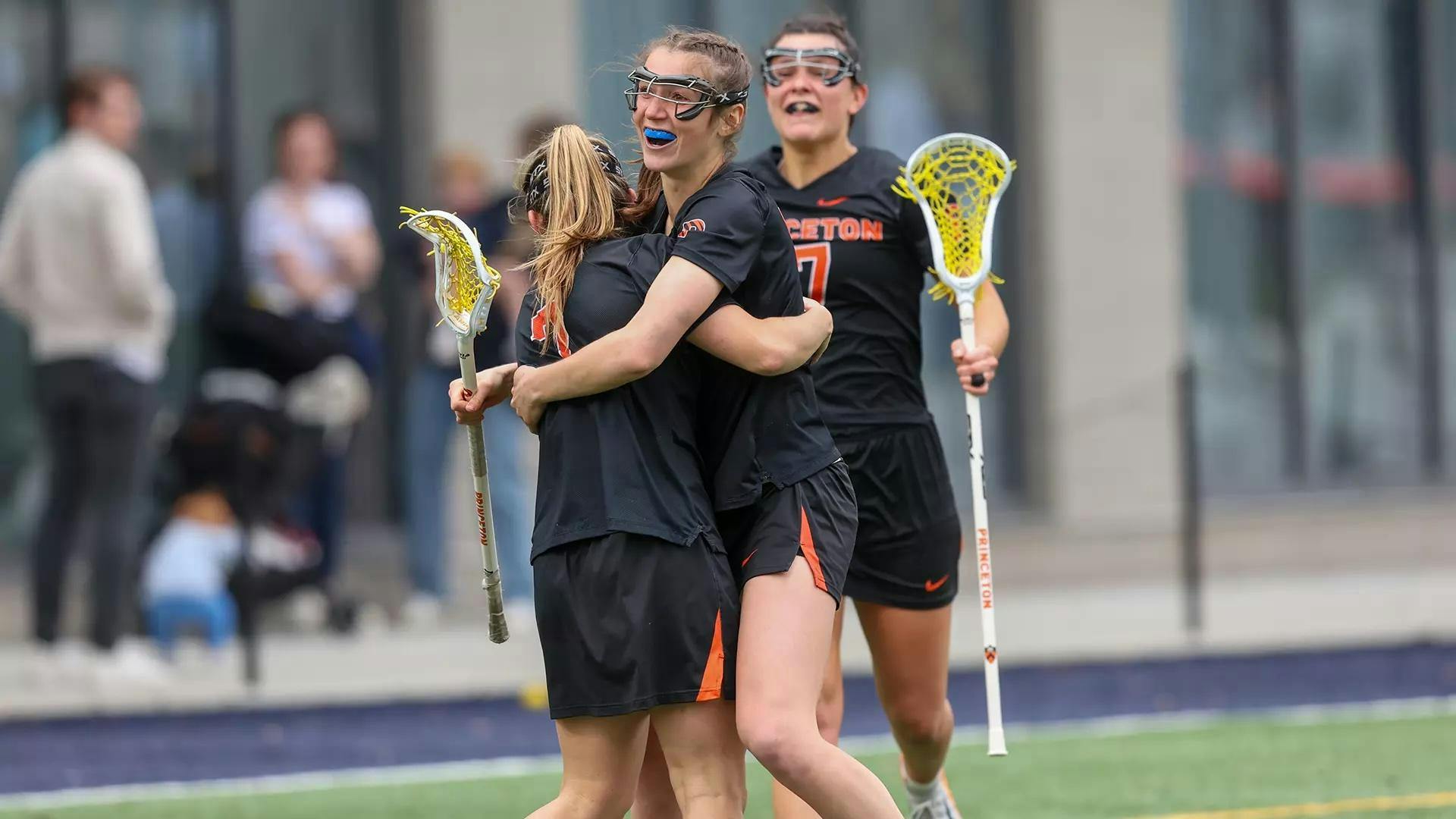 Two women’s lacrosse players in black and orange jerseys embrace as a third jogs in to join them.