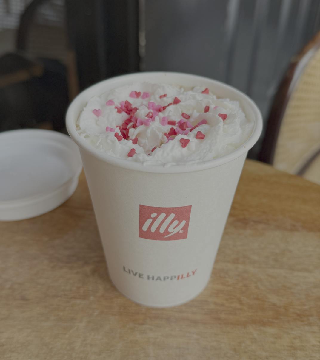A warm drink in a white cup sits on a wooden table, topped with whipped cream and heart-shaped red and pink sprinkles. 