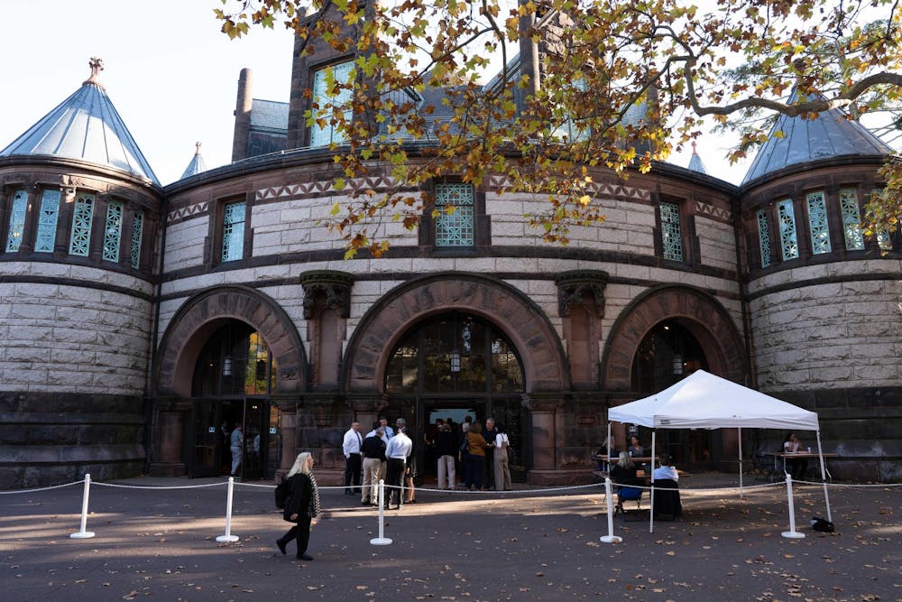 A group of people congregate in front of a stone building. A small white tent with two people sitting under it is visible in the foreground. 