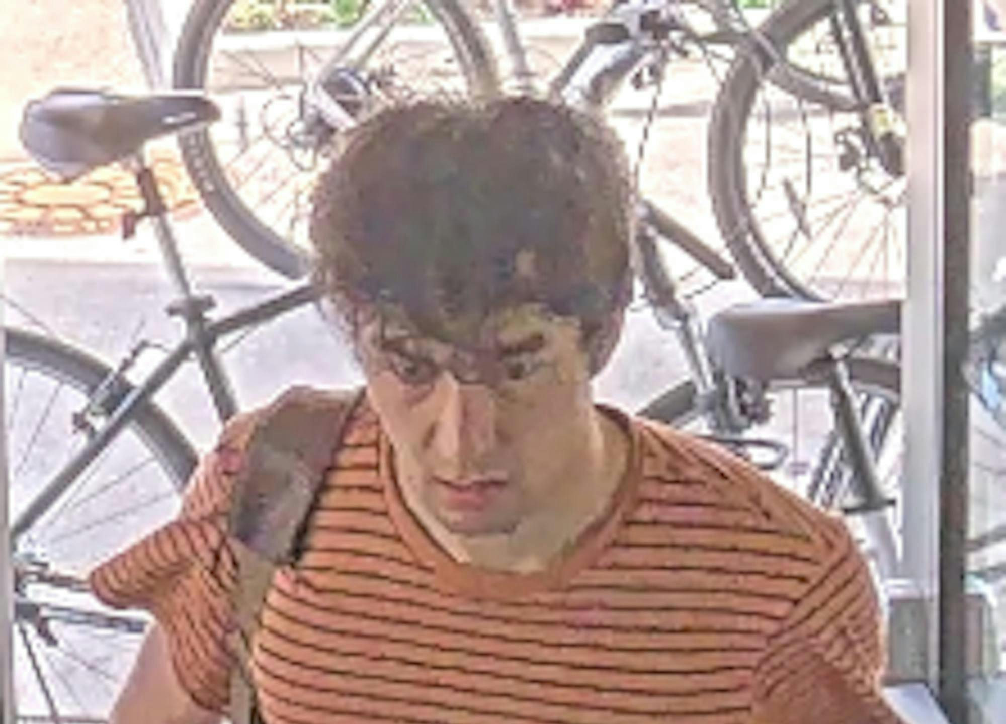 A man with brown hair, orange shirt with black stripes, and backpack stands in front of bikes.