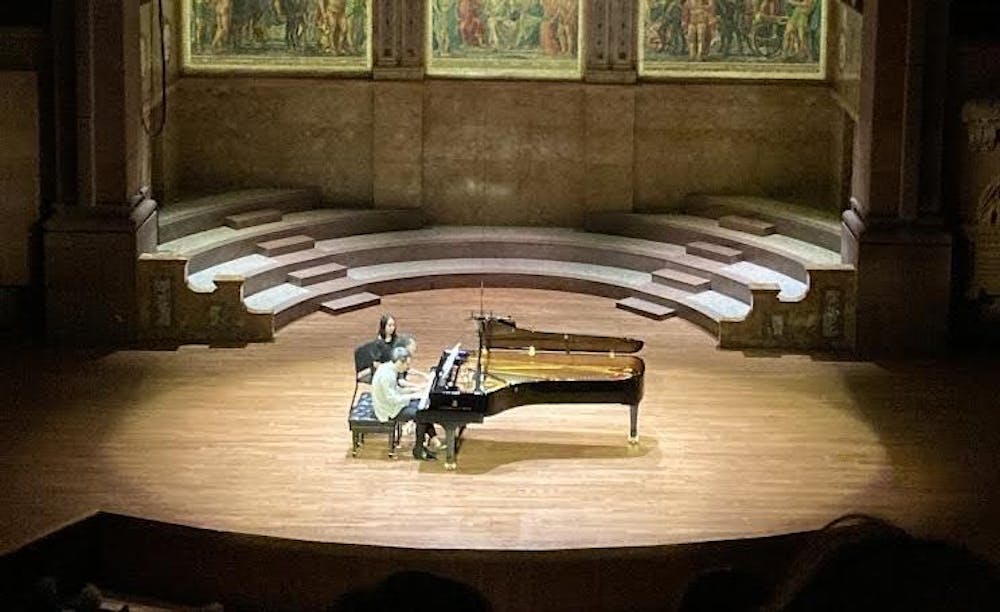 A man and woman sit together at a piano on a stage. The stage light is centered on the two while the rest of the stage is slightly darker.