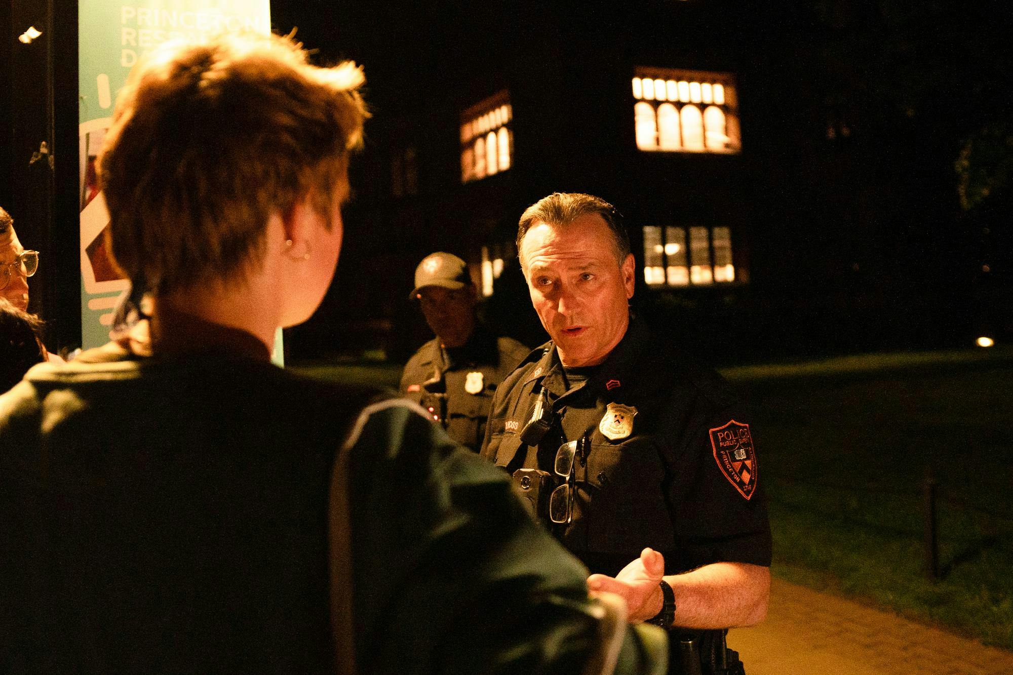 At night, a PSAFE officer in a black and orange uniform interacts with a marshal with orange hair.